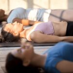 Woman lying on her back in Savasana or Corpse Pose with her arms at her sides, palms up during guided meditation practice.