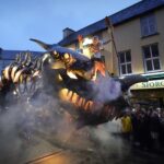 Dramatic smoke plumes as a man stands on top of a moving dragon figure as people watch from below.