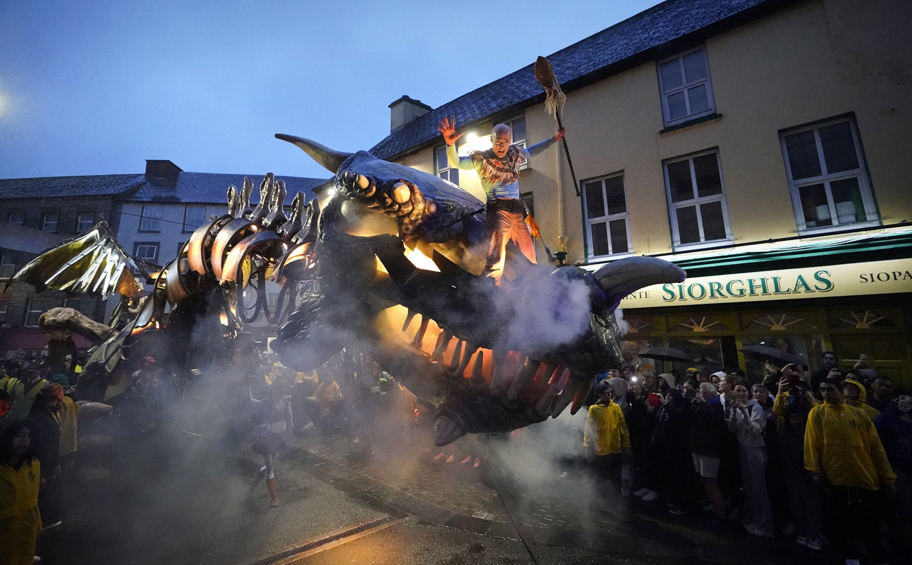 Dramatic smoke plumes as a man stands on top of a moving dragon figure as people watch from below.